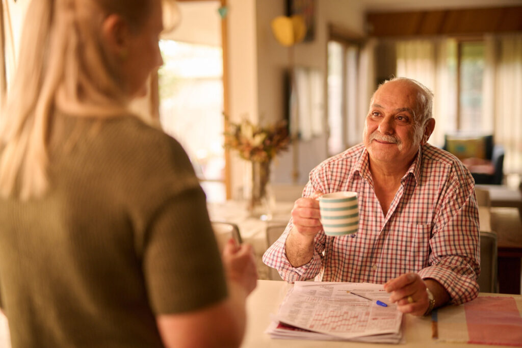 A man and woman chatting at the kitchen table. He is holding a coffee cup.