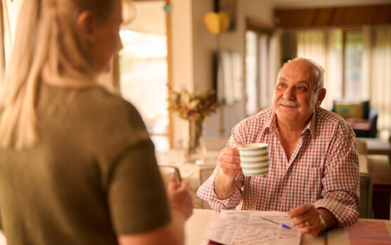 A man and woman chatting at the kitchen table. He is holding a coffee cup.